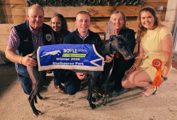 2026 BOYLE Sports Irish Greyhound Derby champion Bockos Gold with the Holland family, trainer Graham Holland, daughter Rachel, son Timmy, wife Nicky and Timmy's partner Sheena. Image shows a family, the Holland family, in front a timber backdrop and kneeling behind a black greyhound, their 2026 BOYLE Sports Irish Greyhound Derby champion, Bockos Gold