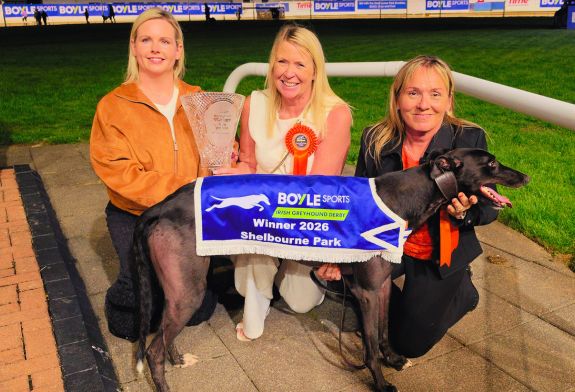 Jenna Boyle, President of BOYLE Sports, hands the winner's trophy to owner Beverly Lochead as they and Nicky Holland, wife of trainer Graham Holland, kneel behind Bockos Gold, 2026 BOYLE Sports Irish Greyhound Derby champion Image shows 3 women, Jenna Boyle, Bev Lochead and Nicky Holland, in the infield in Shelbourne Park kneeling behind a black greyhound, Bockos Gold, wearing the 2026 BOYLE Sports Irish Greyhound Derby winner's jacket. Jenna is handing a large pece of crystal, the winner's trophy, to Bev.