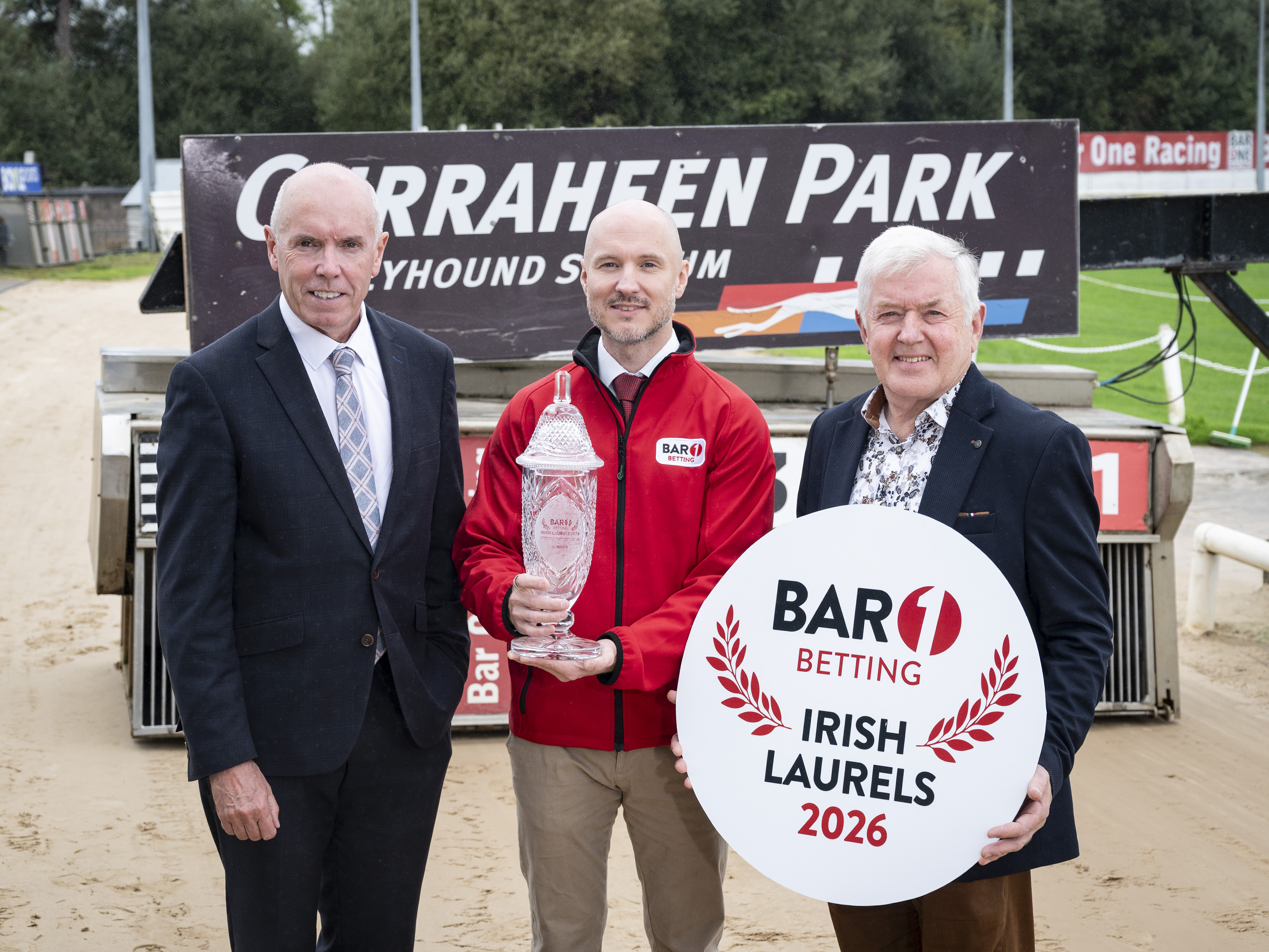 Tim Lucey, CEO of Greyhound Racing Ireland, Aaron Hughes Bar 1 Betting and Pat Flanagan, Chairman of Greyhound Racing Ireland, launching the 2026 Bar 1 Betting Irish Laurels in Curraheen Park The image shows 3 men standing on the sand track at Curraheen Park, the man in the middle is holding a commerative crystal trophy and the man on the right is holding a circular disc with the logo for the 2026 Bar 1 Betting Irish Laurels in Curraheen Park