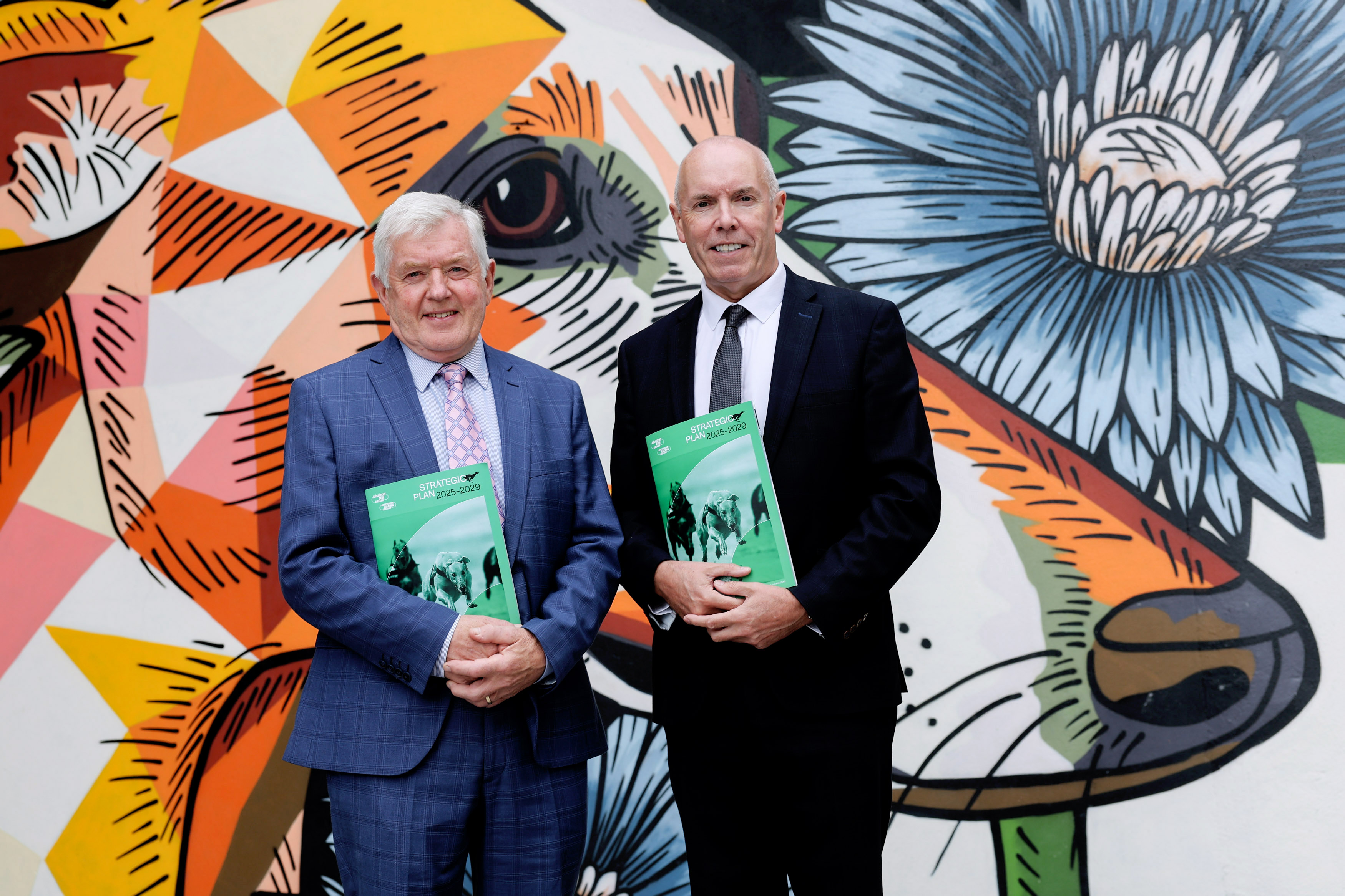 Picture shows two men, RCÉ/GRI Chairman Pat Flanagan and CEO Tim Lucey, standing in front of  colourful mural at Shelbourne Park Greyhound Stadium to launch the new Strategic Plan 2025 - 2029 which they are both holding in their hands.